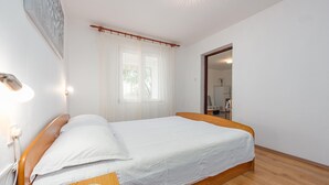 Ein Schlafzimmer mit einem hölzernen Bett, weißen Bettwäsche, einer Nachttischlampe, einem Fenster mit durchsichtigen Vorhängen und Blick in die Küche.