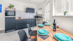 Una cocina moderna con armarios oscuros, un microondas y un lavamanos. Hay una zona de comedor con una mesa de madera preparada para dos personas, con platos turquesa y una botella de vino.