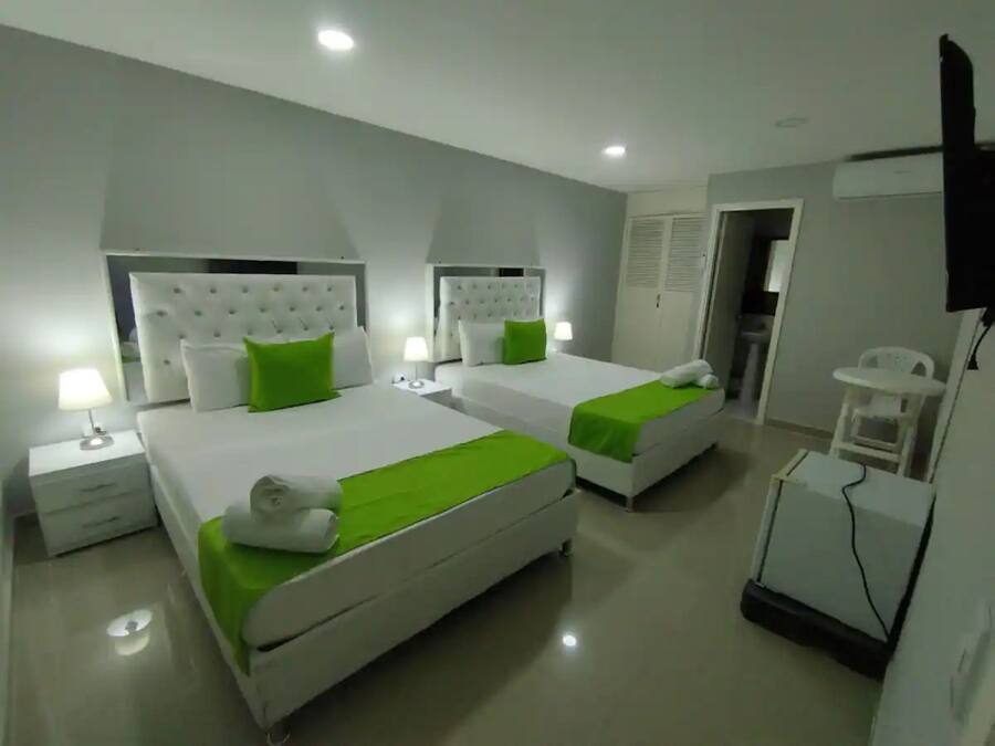 Habitación de hotel con dos camas, cada una con cubrecama verde y cabeceros blancos. Hay mesitas de noche con lámparas, una mesita pequeña con una silla y un televisor montado en la pared.