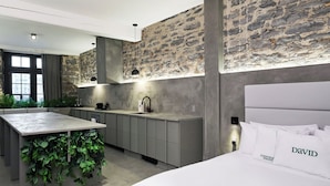 A modern kitchen with a stone wall backsplash, a central island with greenery, and a bed with white bedding and pillows labeled "DAVID". - PREMIUM LOFT | The Signature (Montréal)