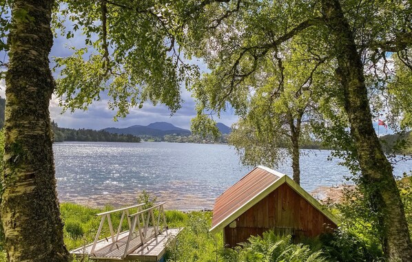 A wooden cabin with a sloped roof, surrounded by trees and overlooking a calm lake with mountains in the distance. - 4 bedroom beach front home in Skodje (Skodje)
