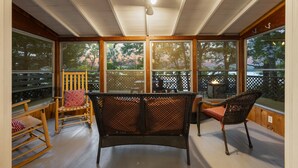 A covered porch with wooden flooring, wicker furniture, and large windows offering views of trees and a deck area. - Seneca Lake Waterfront Cottage with Hot Tub on FLX Wine Trail (Burdett)