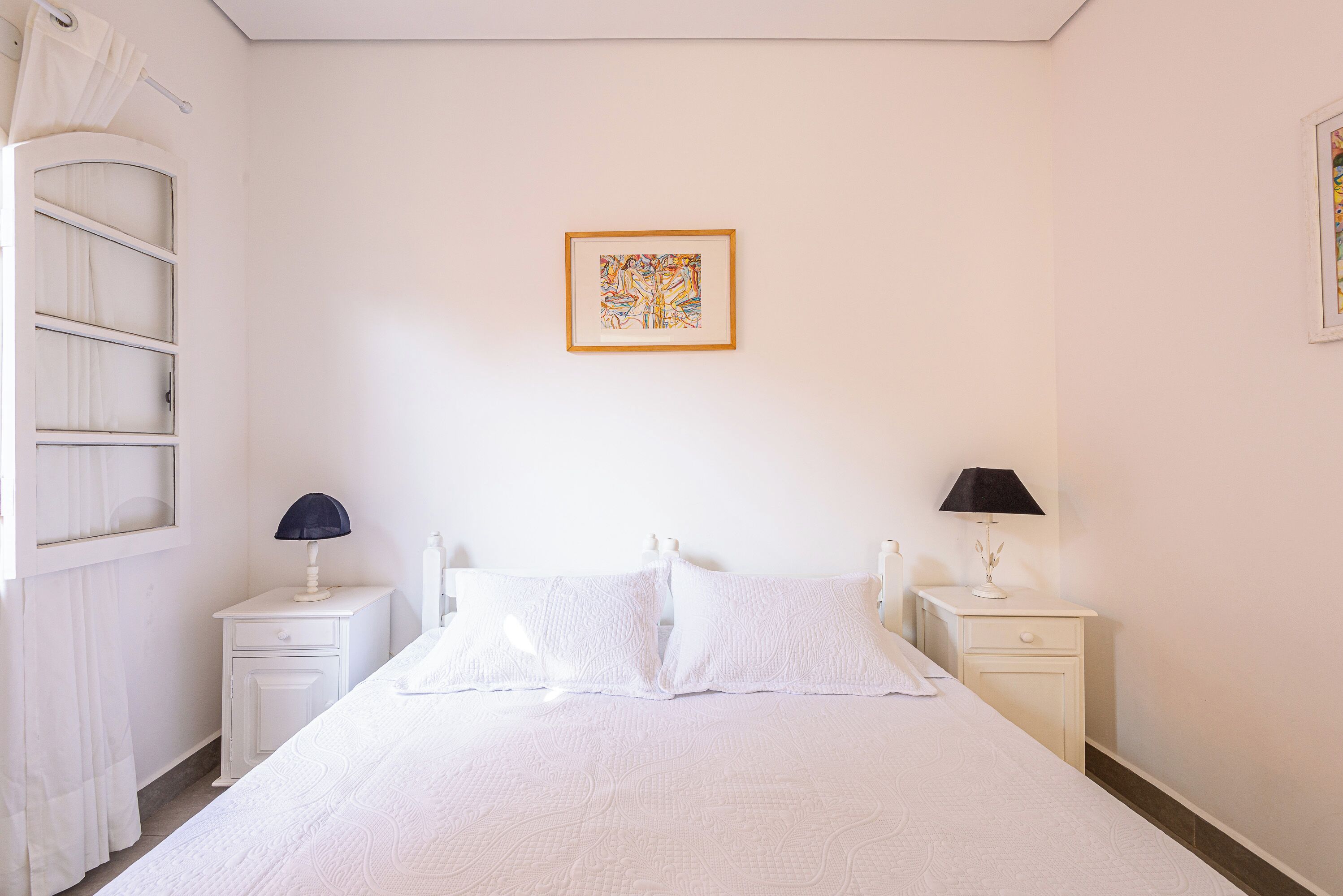 Quarto com uma cama, duas mesas de cabeceira, um quadro na parede e uma janela com cortinas.