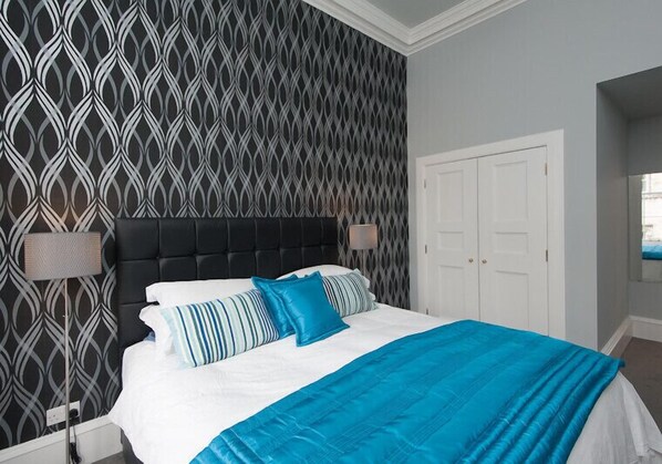 Une chambre avec un papier peint à motifs, une tête de lit noire en tissu, une porte blanche et un lit recouvert d’une couverture bleue et de coussins à rayures.