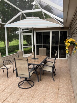 Une terrasse couverte avec une table et des chaises, une jardinière suspendue ornée de fleurs jaunes et une vue sur la verdure environnante.