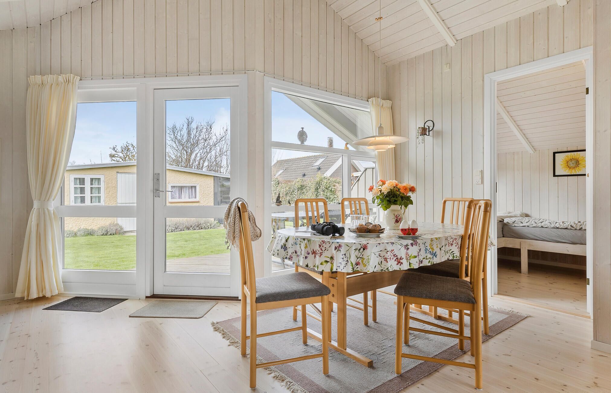 A dining area with a table set for two, wooden chairs, a floral tablecloth, and a vase of flowers.