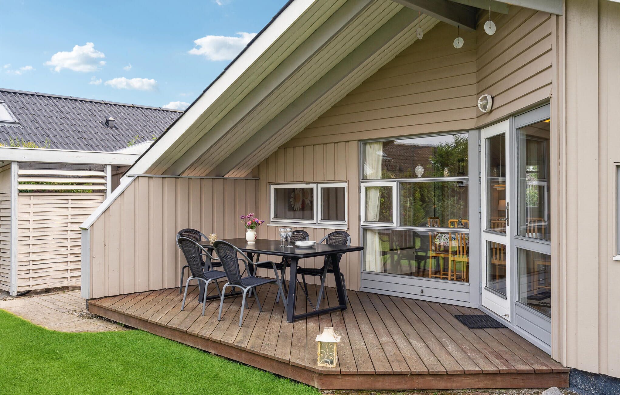 En overdækket terrasse med et bord, stole, en lanterne og en skydedør til en have.