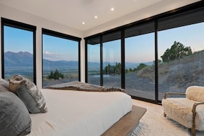 A bedroom with a large bed, a chair, and a view of a mountainous landscape through large windows.