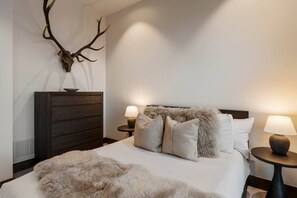 A bedroom with a large bed, two bedside tables with lamps, a wooden dresser, and a wall-mounted deer antler decoration.