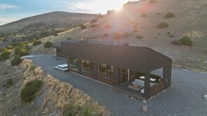 A modern, single-story building with large glass windows and a flat roof, situated on a gravel driveway with a view of a hilly landscape.