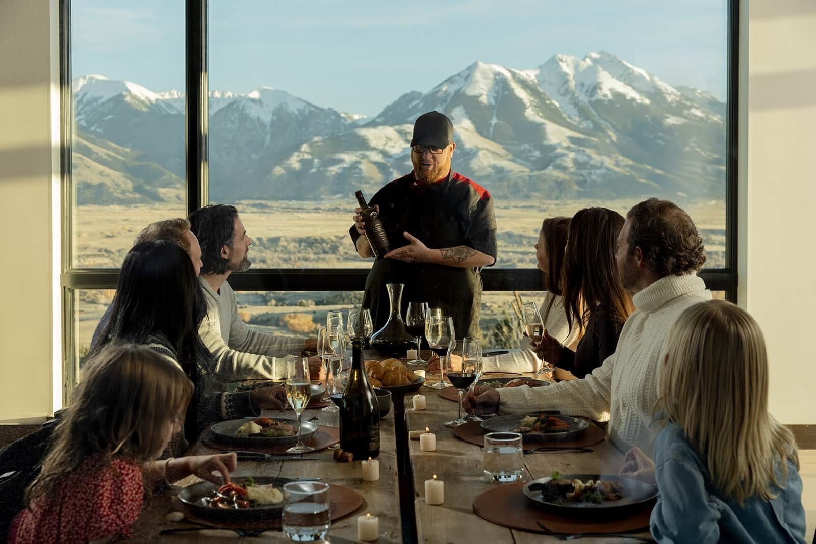 Un groupe de personnes est assis à une table, devant un paysage de montagne, avec une bouteille de vin et des bougies sur la table.