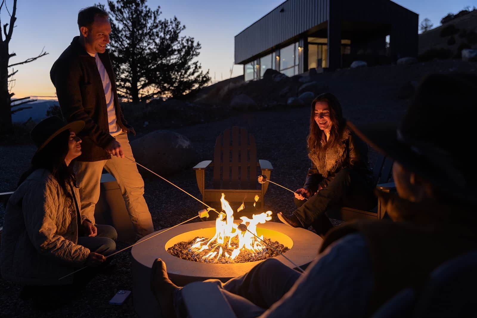 A group of people gathered around a fire pit in an outdoor setting with a modern building in the background.