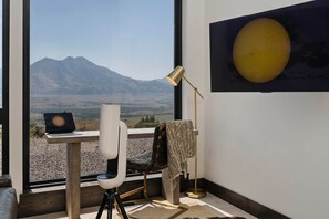 Una habitación con un ventanal grande, un escritorio con una computadora portátil, una silla, una lámpara de pie y un objeto esférico sujeto a la pared.