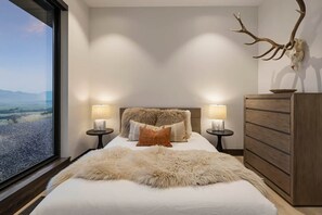 A bedroom with a large window, a bed with a fur throw, a wooden dresser, and a mounted deer skull.