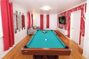 Una mesa de billar con una bola blanca, cortinas rojas y un sofá en una habitación con paredes blancas y un diseño de árboles rojos en una pared.
