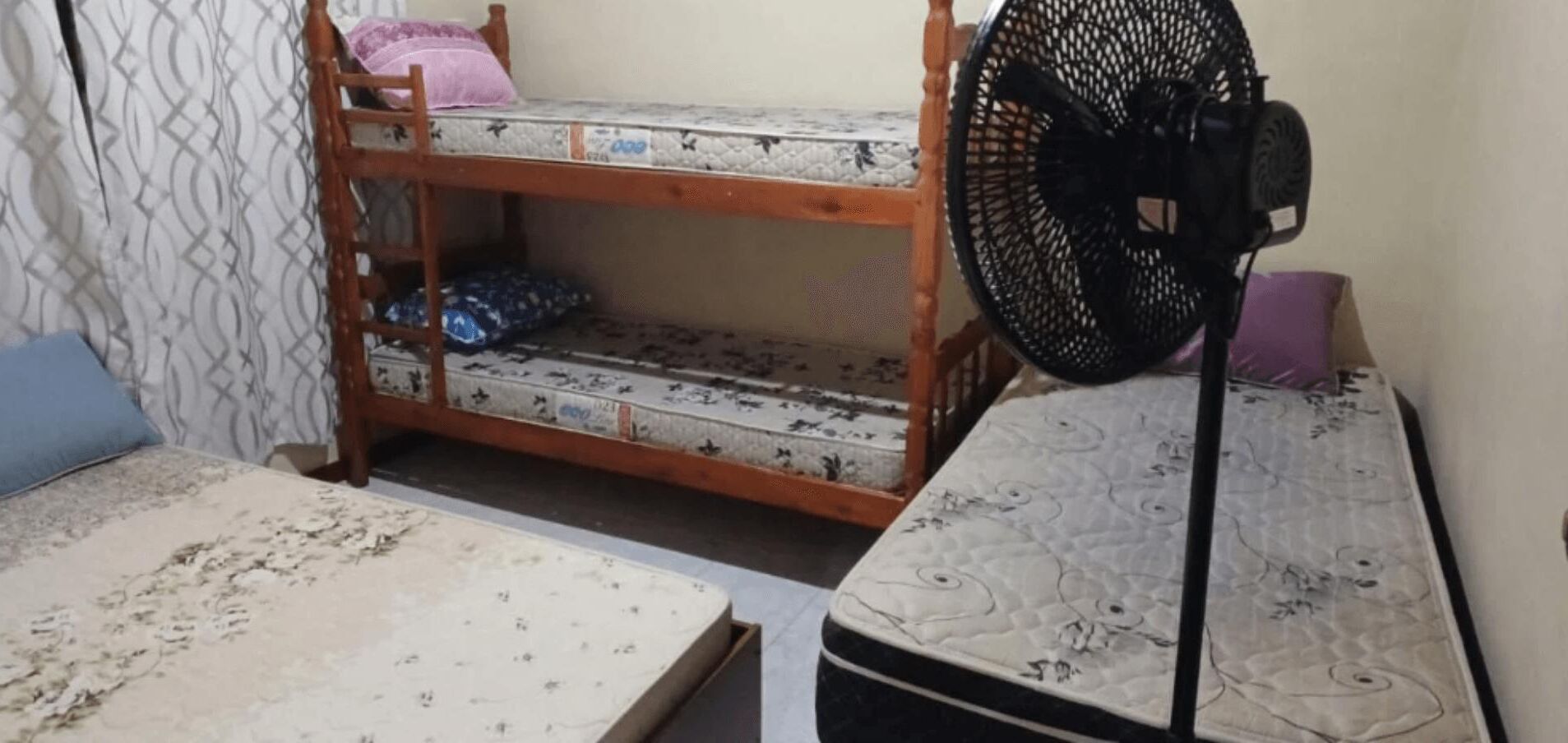 Ein Zimmer mit Etagenbetten, einem Standventilator und einer Matratze auf dem Boden.