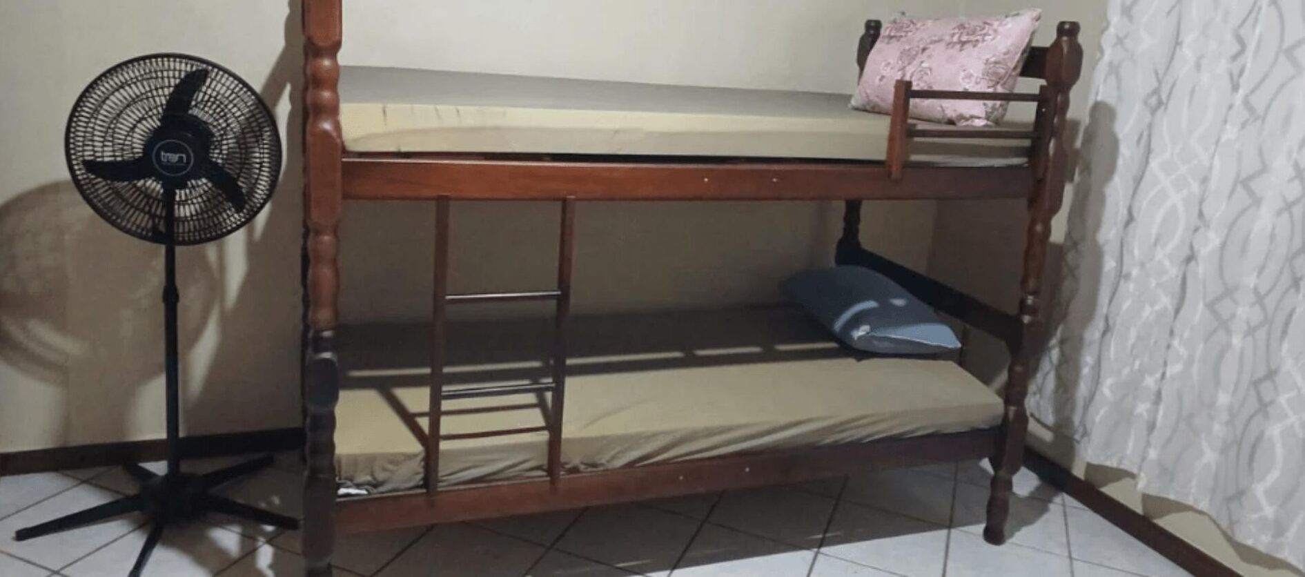A bunk bed with a mattress and a pillow on the top bunk, a fan next to it, and a chair with a cushion.