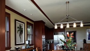 A spacious dining area with a wooden table and chairs, a chandelier, and a kitchen in the background. - Kolea 10D: 3 Bed/3 Bath Garden Level Condo w Direct Access to Pool & Ocean. (Waikoloa Village)