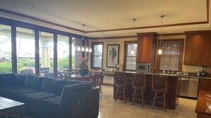 A spacious living area with a dark-colored sofa, a dining table, and a kitchen area with wooden cabinets and a stainless steel refrigerator. - Kolea 10D: 3 Bed/3 Bath Garden Level Condo w Direct Access to Pool & Ocean. (Waikoloa Village)