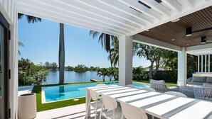 A modern outdoor patio with a pool, white furniture, and a view of a lake. - Modern Waterfront Paradise with Heated Pool - Near the Beach & Aventura Mall! (Miami)