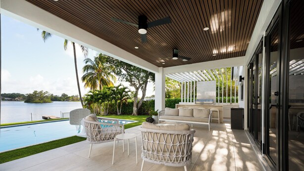 A modern outdoor living area with a pool, white wicker furniture, and a view of a lake. - Modern Waterfront Paradise with Heated Pool - Near the Beach & Aventura Mall! (Miami)