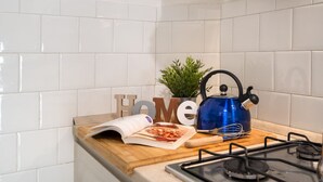 Uma cozinha com azulejos brancos, uma chaleira azul, uma tábua de corte de madeira, uma planta e uma revista.