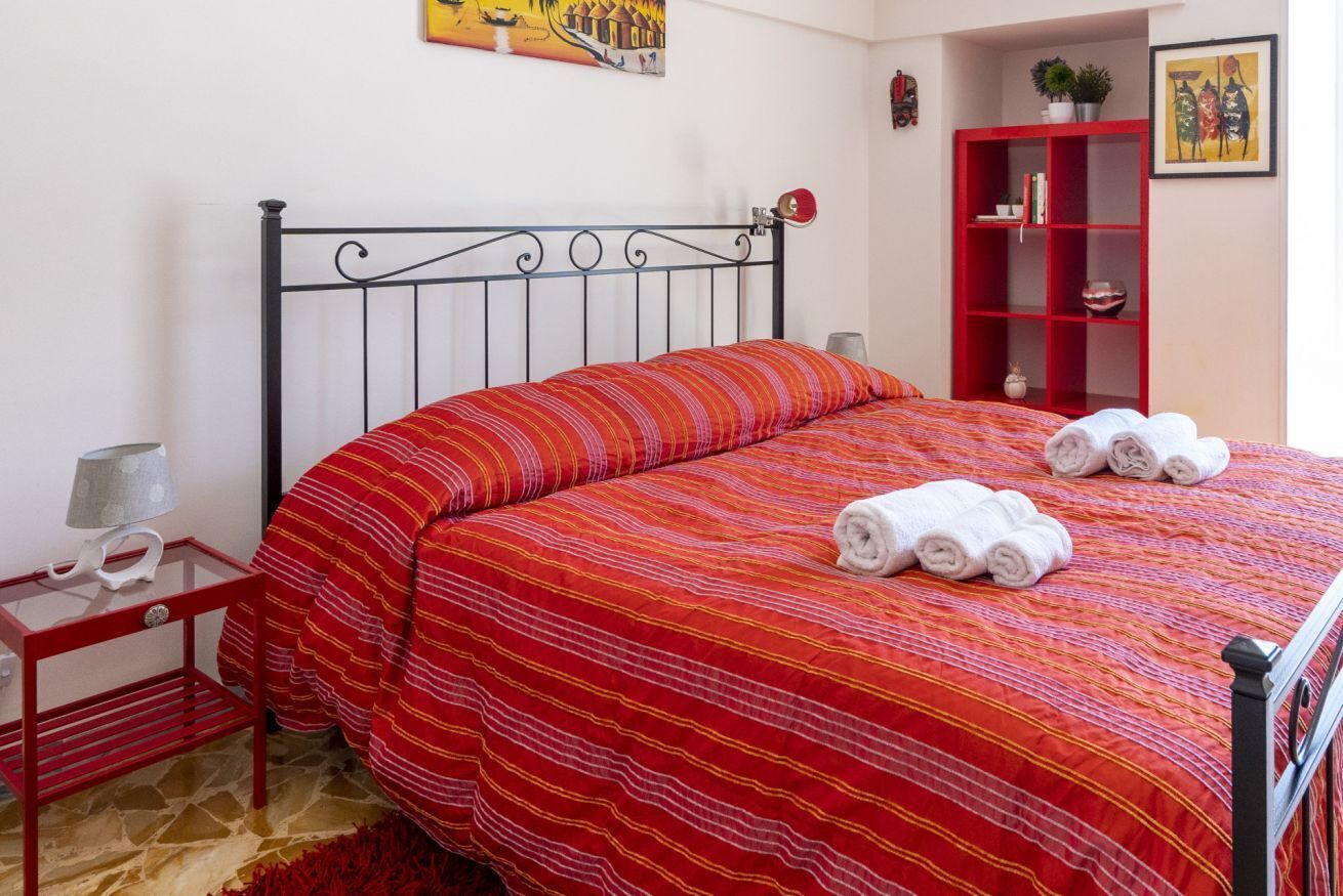 A neatly made bed with a striped red and yellow bedspread, a red nightstand with a lamp, and a red bookshelf with books and decorative items.