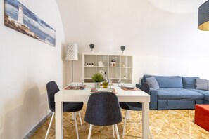 Un comedor con una mesa blanca, cuatro sillas, un sofá azul, una estantería y un cuadro de un faro.