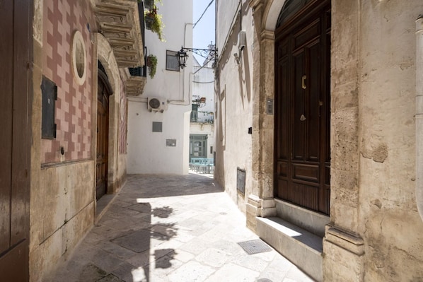 A narrow alleyway with stone pavement, a wooden door, and a white building with a checkered pattern.