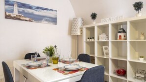Uma sala de jantar com mesa branca posta para quatro pessoas, uma pintura de um farol, uma estante branca com objetos decorativos e um abajur com cúpula listrada.