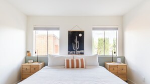 Ein Schlafzimmer mit einem großen Bett, zwei Nachttischen, einem Wandbild und Fenstern mit Blick auf Gebäude und Bäume.
