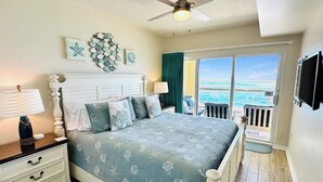 Ein Schlafzimmer mit einem Bett, einem Nachttisch, einer Kommode, einem Deckenventilator und Blick auf den Ozean durch eine Schiebetür.