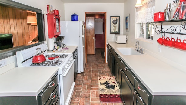 A compact kitchen with a stove, sink, and refrigerator. A red kettle and a blue container are on the counter.