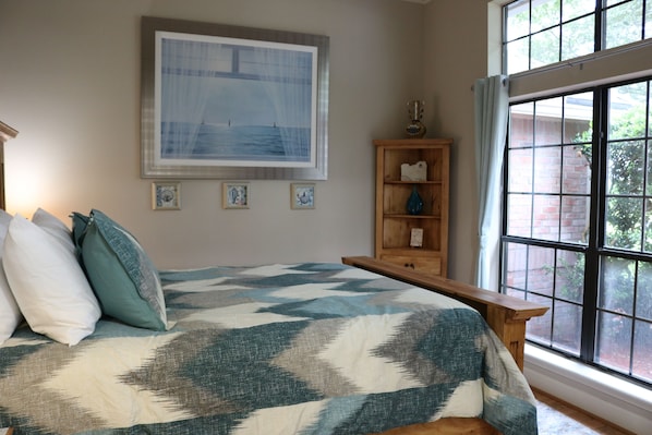 Quarto com cama grande, mesa de cabeceira de madeira, janela com vista para área verde e quadro na parede.