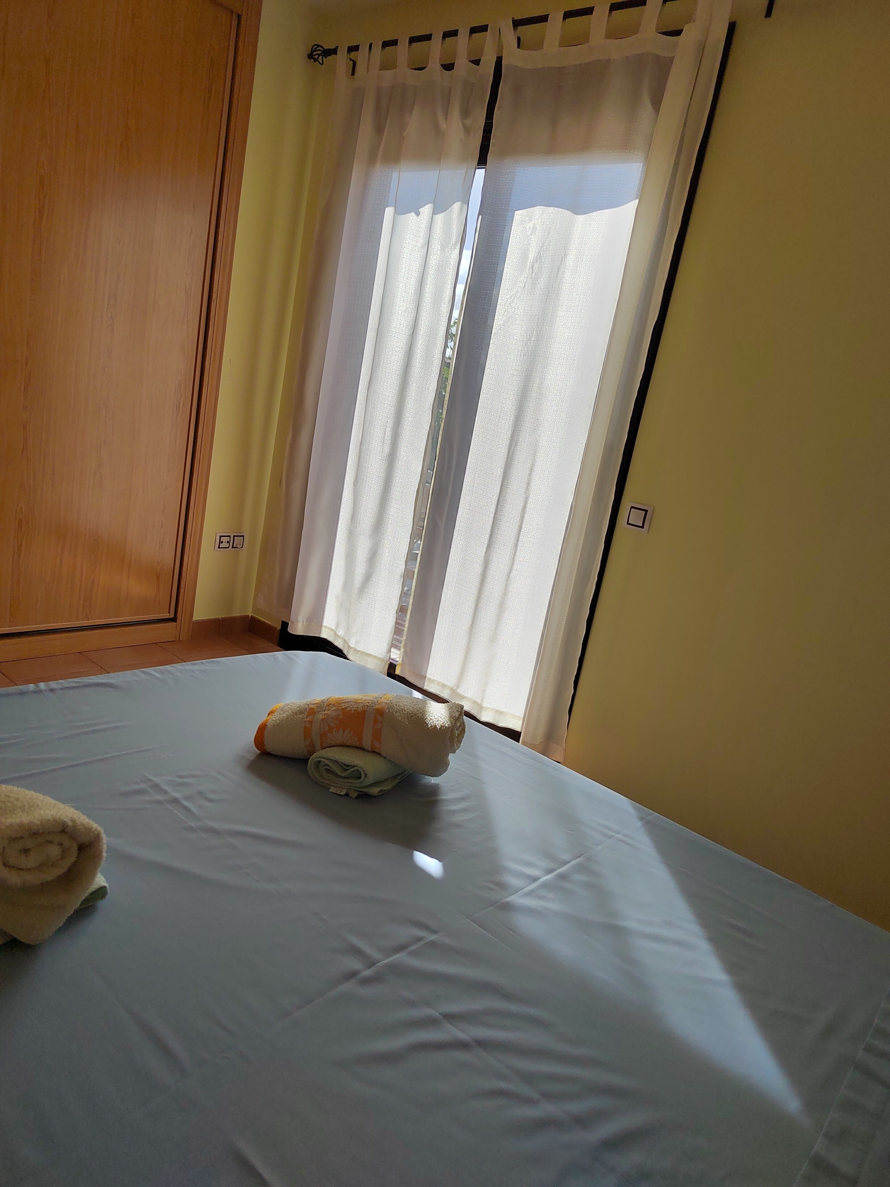 Una cama bien hecha con una toalla enrollada y una almohada, un armario de madera y una ventana con cortinas translúcidas.