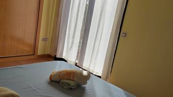 Una cama bien hecha con una toalla enrollada y una almohada, un armario de madera y una ventana con cortinas translúcidas.