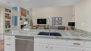 A kitchen with a granite countertop, white cabinetry, and a dishwasher. There is a dining area with a sofa and chairs, and a TV mounted on the wall.