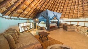 Een knus ingerichte kamer met rieten dak, een bed met beddengoed met dessin, een klein tafeltje en een comfortabele bank.