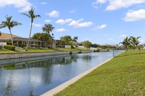 A canal with a grassy bank, palm trees, and buildings in the background.