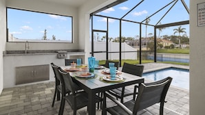 Un área de comedor al aire libre con vista a la piscina, una parrilla y un ventanal grande.