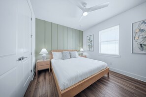 Un dormitori amb llit, taules de nit, ventilador de sostre, finestra amb persianes i una obra d'art penjada a la paret.
