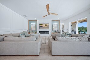 Een moderne woonkamer met twee grijze banken, een plafondventilator, een flatscreen-televisie en een groot raam met uitzicht op de omgeving.