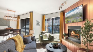 A modern living room with a sofa, coffee table, fireplace, TV, and a view of the outdoors through large windows.