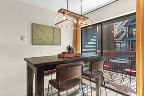 Un coin repas avec une table en bois foncé, deux chaises marron, un luminaire suspendu et une vue sur un balcon agrémenté de chaises rouges et d’un tapis à motifs.