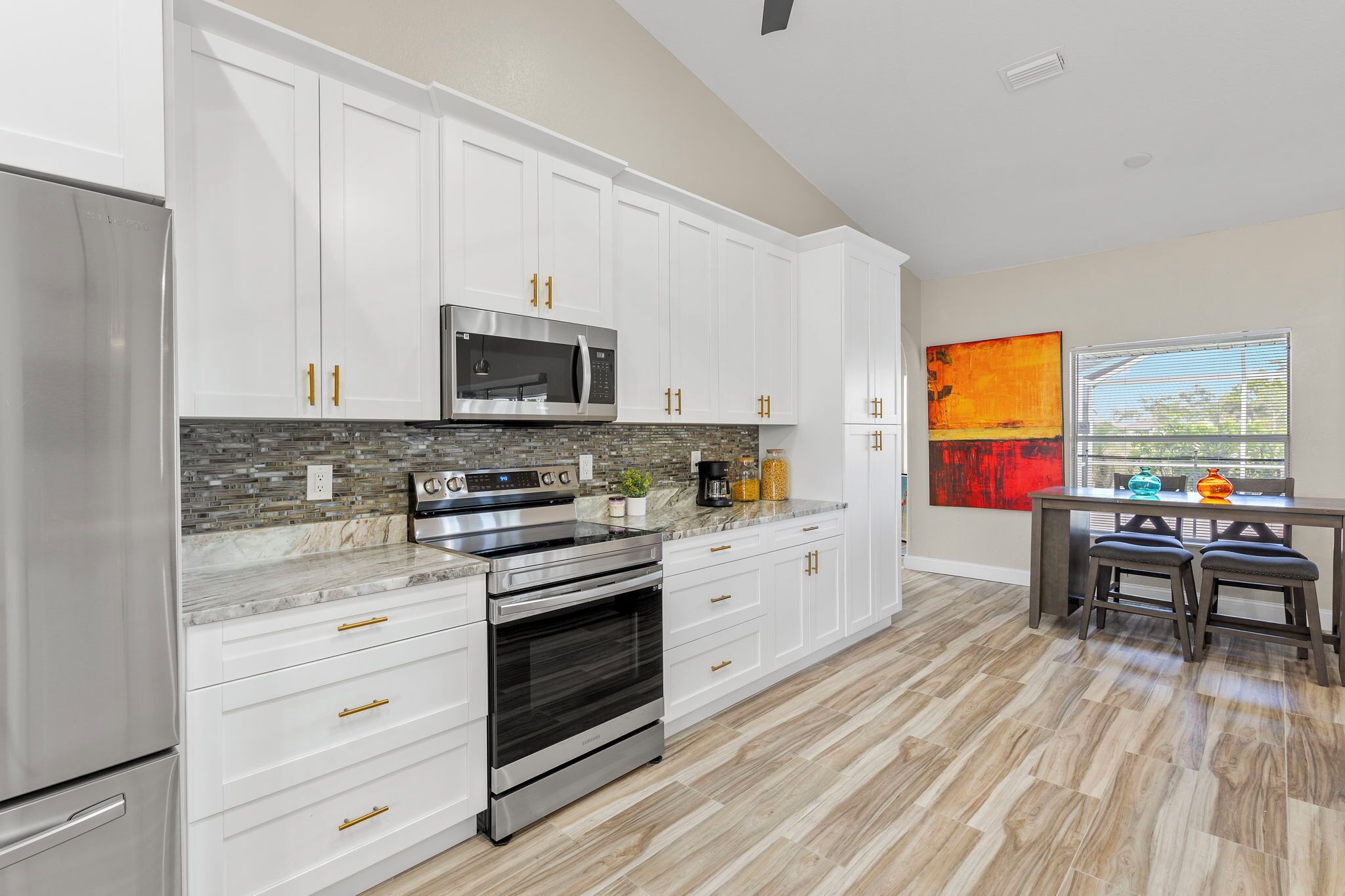 A modern kitchen with white cabinets, a stainless steel refrigerator, a microwave, and an oven. There is a dining table with chairs and a window with blinds.