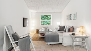 A modern, minimalist living room with a white sofa, a small white table, and a chair. There is a large window with a view of greenery outside. - 3 bedroom pet friendly home in Græsted (Græsted)