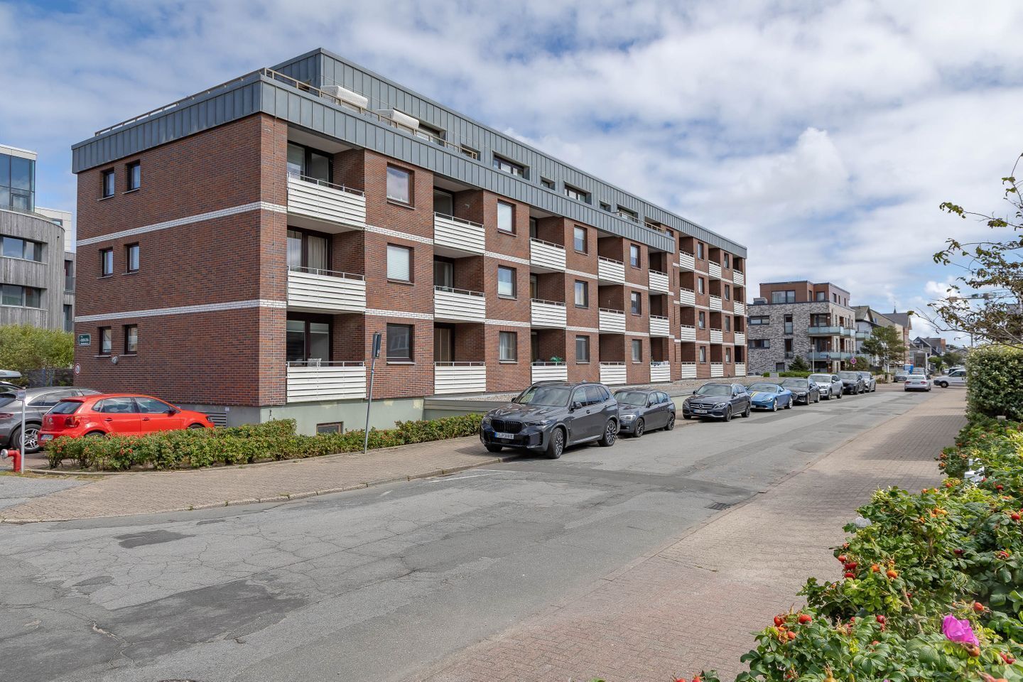 A multi-story residential building with a brick facade, balconies, and a parking area with several cars.