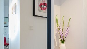 Un pasillo con pared blanca, una obra de arte enmarcada de labios y un jarrón blanco con flores moradas sobre un estante de madera.