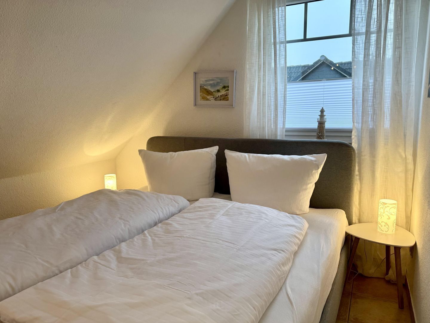 Ein ordentlich bezogenes Bett mit weißer Bettwäsche und Kissen, ein kleiner Nachttisch mit einer Lampe und ein Fenster mit durchsichtigen Vorhängen.