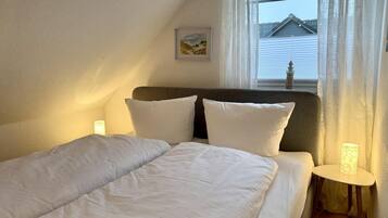 Ein ordentlich bezogenes Bett mit weißer Bettwäsche und Kissen, ein kleiner Nachttisch mit einer Lampe und ein Fenster mit durchsichtigen Vorhängen.